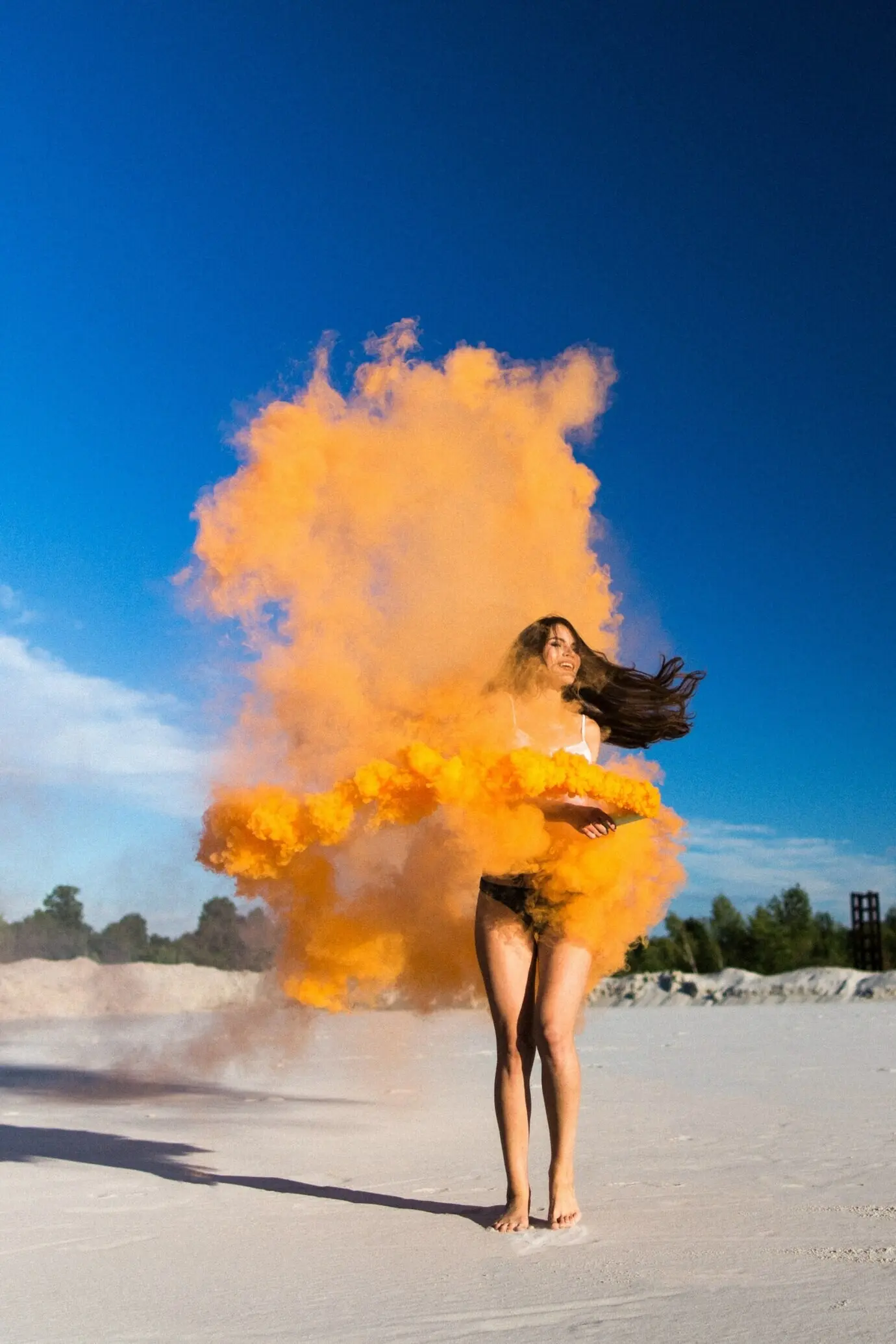 Frau tanzt mit orangem Rauch an weißem Strand unter blauem Himmel.